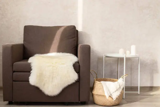A sheep skin on a chair, next to a wicker basket with a blanket and a coffee table with candles. Minimalist interior of the living room. Copy space.
