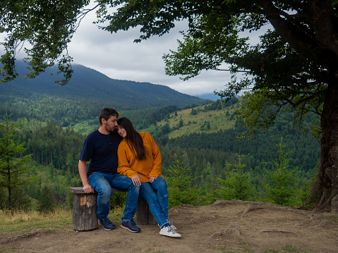 Young man and woman admiring breathtaking view while sitting on bench in the mountains. Lovely couple in casual clothes sit under large tree with forest background.