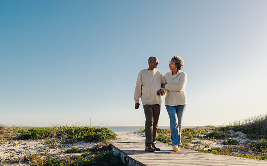 Romantic elderly couple smiling cheerfully while walking down a wooden foot bridge at the beach. Happy senior couple enjoying a refreshing holiday after retirement.