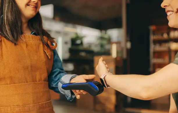 Happy customer scanning her smartwatch on a credit card machine to pay her bill in a cafe. Cheerful woman doing a cashless and contactless transaction using NFC technology.