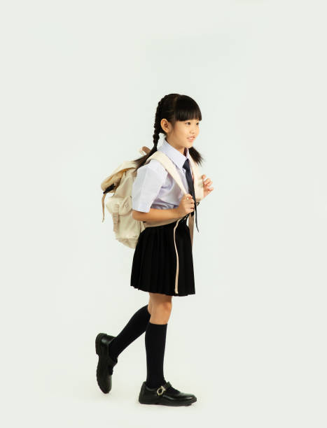 istock Back to school. Asian junior school student in british international uniform carrying backpack walking white background. 1431298225