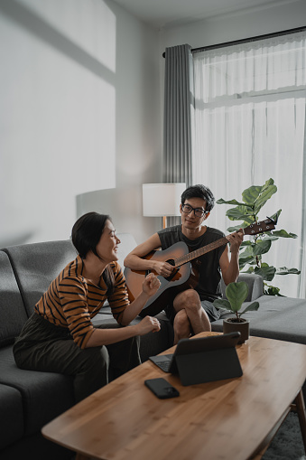 Asian couple play music on guitar together while sitting on comfortable couch in living room enjoying weekend at home.