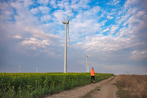 Jogging on dirt road by the agricultural field with wind turbines under the cloudy blue sky, cross-country running
