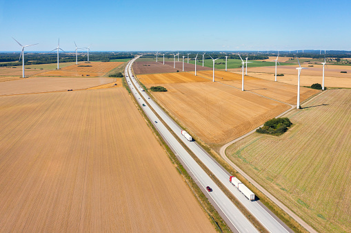 Semi-trucks with trailers on a highway through an agricultural landscape with wind turbines in the Halland region of Sweden.