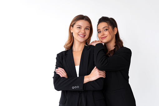Concept of partnership in business. Two Young businesswoman dressed black formal suit standing studio isolated white background. Brunette woman put her hands on the shoulders of her friend.