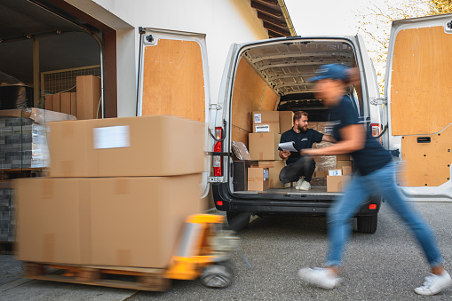 Focus on young delivery man sitting in cargo space of the van, surrounded by boxes, delivery woman rushing and pushing forklift in foreground, blurred.