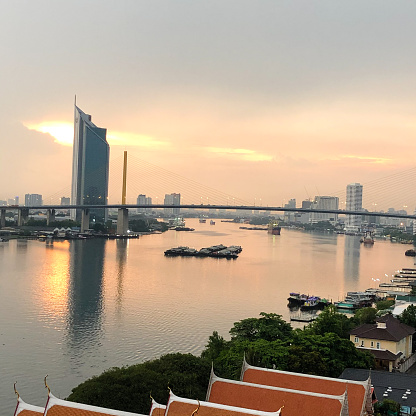 A Bangkok cityscape, the reflection of sunset in The Chao Phraya River with a long suspension bridges and skyscrapers along the waterfront (the orange temple roof is foreground) with cloudy and sunshine evening in square view.
