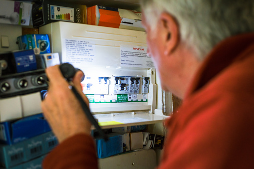 A senior man investigates his fuse box at home - by the light of a battery-operated torch light - in a blackout during the energy crisis.