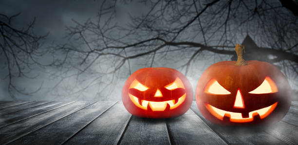 istock Two spooky Halloween pumpkins, Jack O Lantern, with an evil faces and eyes on a wooden table with a misty gray background. 1431347624