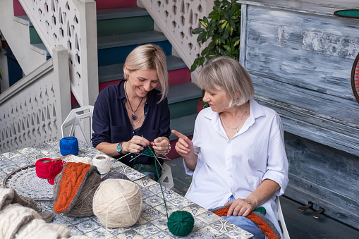 Mom and daughter knit together in the garden. Family friendly leisure. Elderly mother teaching to crochet adult daughter