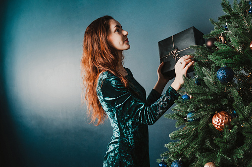 Beautiful girl with a gift box by the Christmas tree, smiling. New Year and Christmas concept. Home and family warmth. Copy space for your text and design