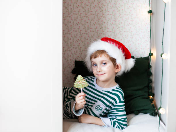 little boy wearing striped pajamas and santa claus hat having fun sitting in a white wardrobe with a lollipop. - christmas child little boys peeking imagens e fotografias de stock