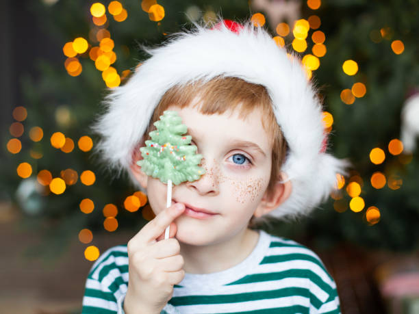 close up of little boy wearing striped pajamas and santa claus hat with a lollipop closing one eye. - christmas child little boys peeking imagens e fotografias de stock