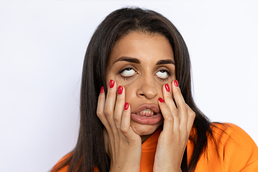 Young Hispanic woman rolling up eyes. Female figure in orange hoodie holding hands on face, rolling up eyes with annoyance, grimacing, making crazy faces. Portrait, studio shot, grimacing concept