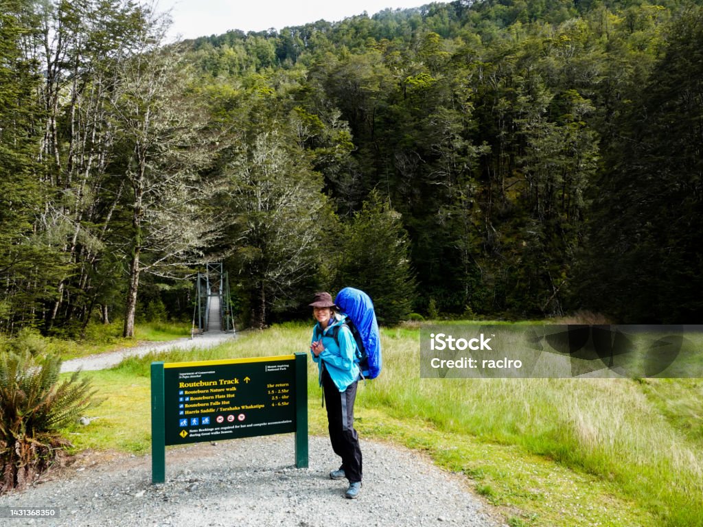Mature woman backpacker standing by route sign on the Glenorchy side ready to start. Routeburn Track, New Zealand. 6-11 Months Stock Photo
