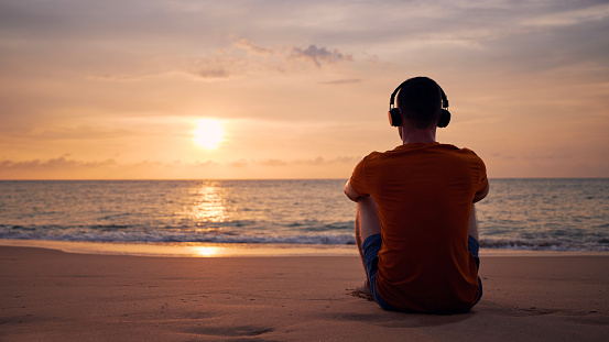 Rear view of man with headphones on beach. Serenity, contemplation and listening music at beautiful sunset.