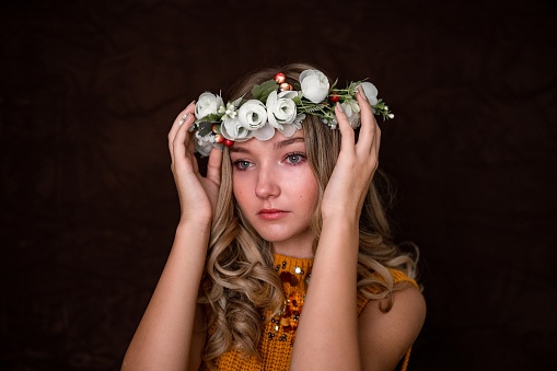 Portrait of a beautiful young girl holding a flower headband on her head while posing in a studio with dark brown background.