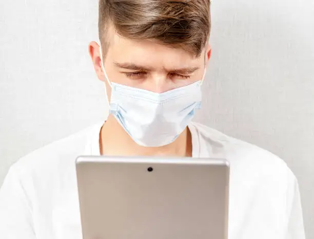Sad Young Man in Flu Mask with a Tablet by the Wall in the Room