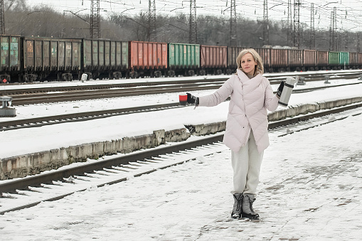 Cheerful active middle-aged woman runs along the platform of the station with a thermos and a mug in the winter season through the snow in warm winter clothes and gloves
