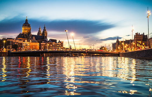 City centre lights in Amsterdam reflected in the water at dusk, with the Basilica of Saint Nicholas on the horizon at left, and the central train station at right.