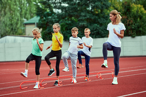 Female coach training athletes. Group of children running on treadmill at the stadium. Concept of sport, achievements, studying, goals, skills. Little boys and girls in sportswear training outdoor.