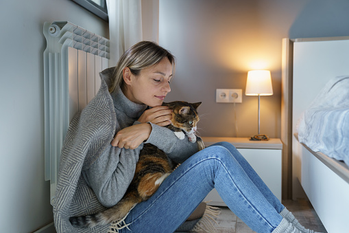 Young woman covered with warm plaid hugs favourite cat. Tired female owner enjoys playing with animal trying to be motivate after losing job. Cat makes blonde lady happy sitting near heating radiator