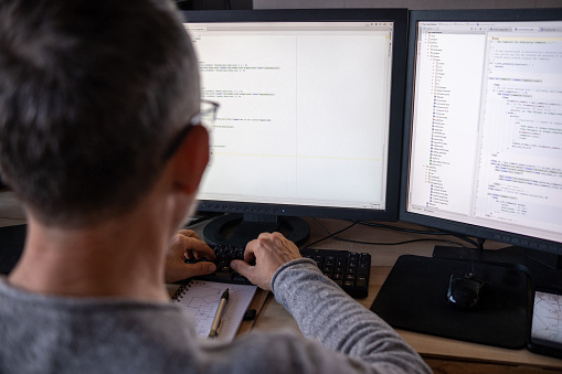 A programmer wearing a gray sweater and glasses writes code. Works on two monitors.  The focus is on the monitor.