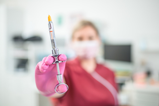An unrecognizable Caucasian female dentist wearing surgical gloves, holding a syringe, preparing oral anesthesia.