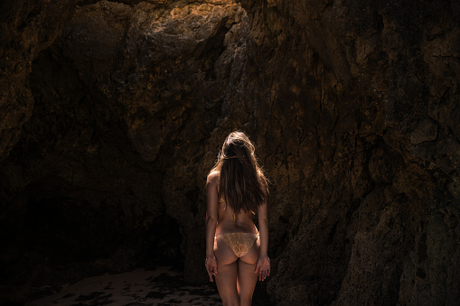 back view of woman in swimwear standing on sandy coast near rocky formation and looking up while enjoying sunny day in Algarve