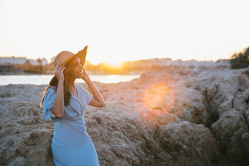 Cheerful young stylish woman tourist in fashionable dress smiling and looking away while leaning on rocky formation under sunset sky and adjusting straw hat during summer holidays in Portugal