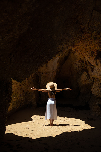Back view of anonymous woman in swimwear and hat standing in sunlit cave with rough stone walls during summer vacation in Algarve, Portugal.