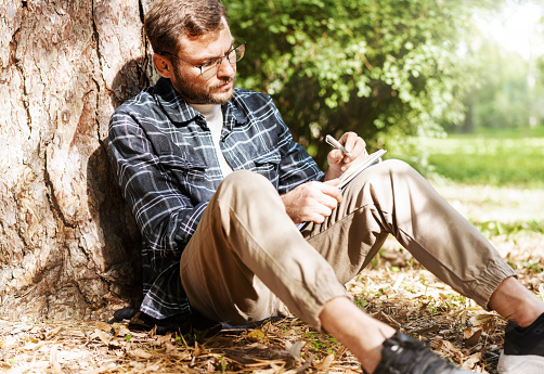 Adult man with eyeglasses sitting in the park and writing in his diary.