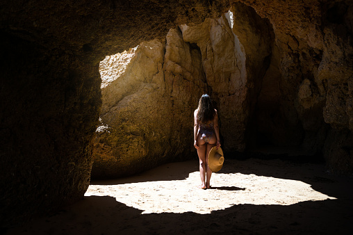 Back view of anonymous woman in swimwear and hat standing in sunlit cave with rough stone walls during summer vacation in Algarve, Portugal.