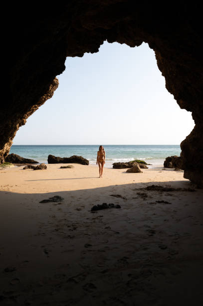 woman in bikini strolling on sandy seacoast near cliffs