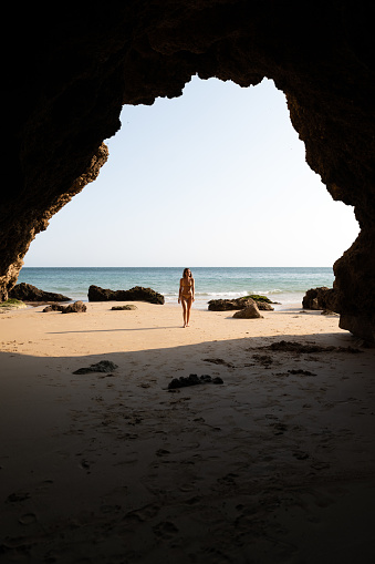 Full body of fit young female traveler in bikini walking on sandy beach near massive rocky cliffs and wavy sea on sunny day in Algarve