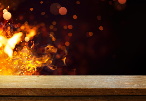 Side view of an empty wooden tabletop with orange fire or flames and sparkles on a dark background.