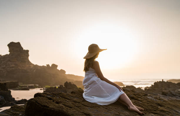 Full body side view of anonymous female traveler in straw hat sitting on stony coast while enjoying sea at sunset time