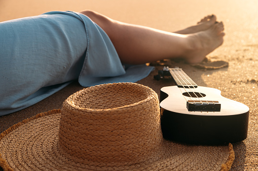 From above of crop anonymous female traveler sitting on sandy beach near straw hat and ukulele on sunny summer day