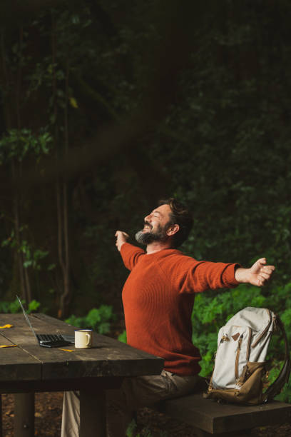 One traveler man outstretching arms after work time in the nature forest using laptop and roaming internet connection. Concept of people and digital life job. Happy adult male with backpack. Green stock photo