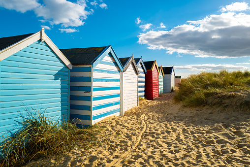 Row of brightly painted beach houses seen on golden sand by sand dunes. Located on the Suffolk coast in the UK. The row of huts extends along the beach,