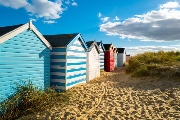 Row of brightly painted beach houses seen on golden sand by sand dunes. Located on the Suffolk coast in the UK. Row of brightly painted beach houses seen on golden sand by sand dunes. Located on the Suffolk coast in the UK. The row of huts extends along the beach, pretty beach house stock pictures, royalty-free photos & images