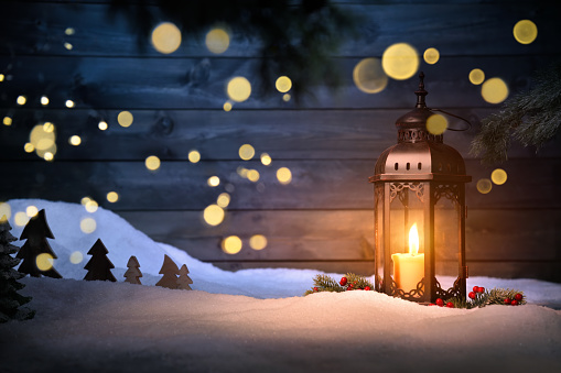 Christmas scene with a lantern, ornaments and blurred lights hanging in front of a dark wooden wall. Warm gold candlelight in contrast with cool blue background
