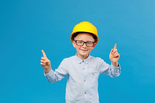 Cute boy construction worker in yellow hard hat standing in indoor studio on blue background and pointing direction with fingers. Future construction worker