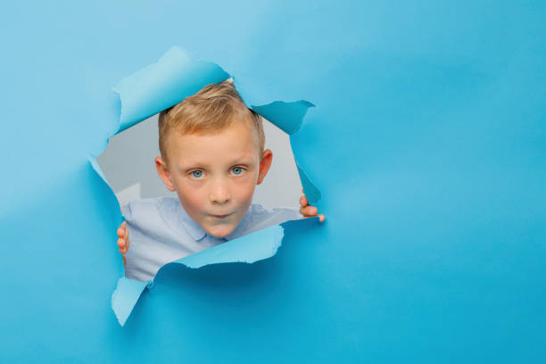 istock Happy cute boy is having fun played on blue background wall, climbs through a hole in the paper. 1431446636