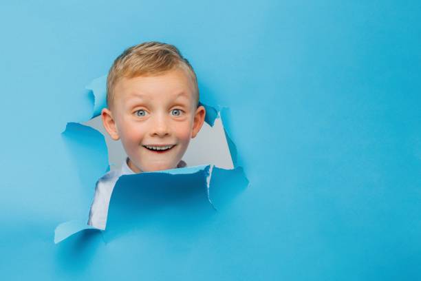 istock Happy cute boy is having fun played on blue background wall, climbs through a hole in the paper. 1431446637