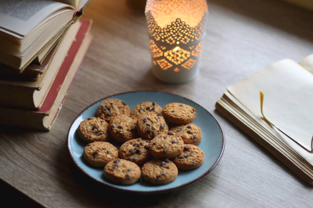 Plate of chocolate chip cookies, stack of vintage books, reading glasses, cup of tea or coffee, lit candle and fairy lights. Hygge at home. Selective focus.