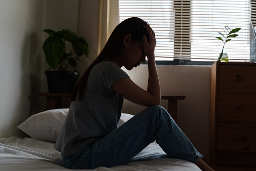 Young Asian woman sitting alone in bedroom, feeling bad and unhappy while thinking about her problems, representing failure, lost love, depression. Dark and moody, negative emotions.