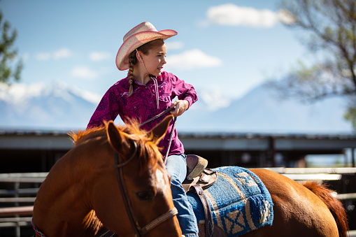 A young, female equestrian donning a pink hat and a pink design button down shirt sitting on a horse inside a ranch during the day.