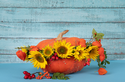 A large pumpkin is decorated with yellow sunflowers. Other berries and fruits also hang on the decoration. The fruit stands on a blue background and the background consists of blue wooden slats.