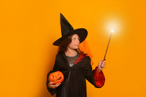 Little cute girl in wizard student costume holding luminous magic wand or glowing magic stick with carved Halloween pumpkin or Jack o lantern pumpkin and looking at light on a yellow background.